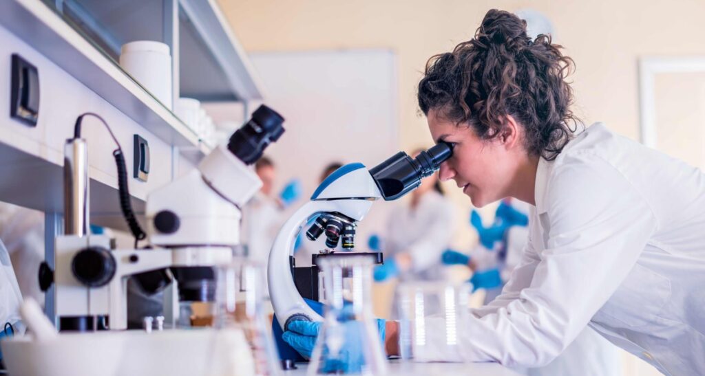 A woman in a white lab coat and wearing blue gloves looks into a microscope. She is working in a laboratory.