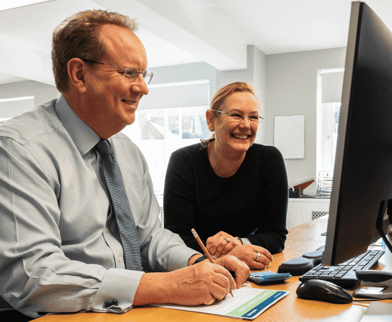 A man and woman in business dress sit at a desk together looking at a computer screen