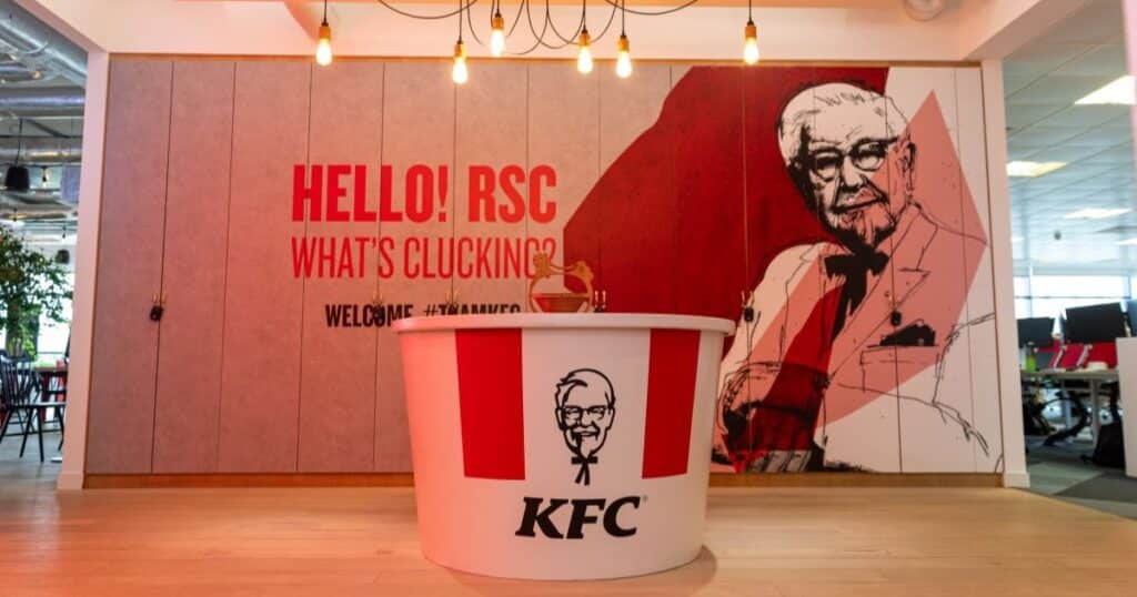 A red and white bucket with the words KFC on it is on a table. In the background is a picture of Colonel Sanders, the founder of KFC.