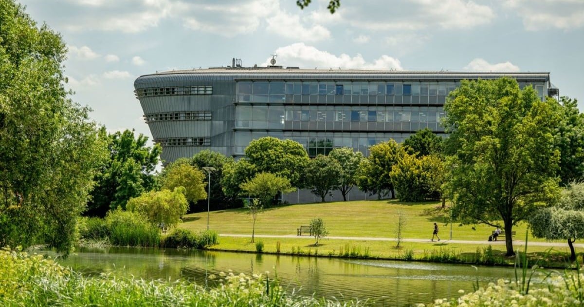 The exterior of a modern glass building standing in leafy surroundings, including trees, lawn and a large lake