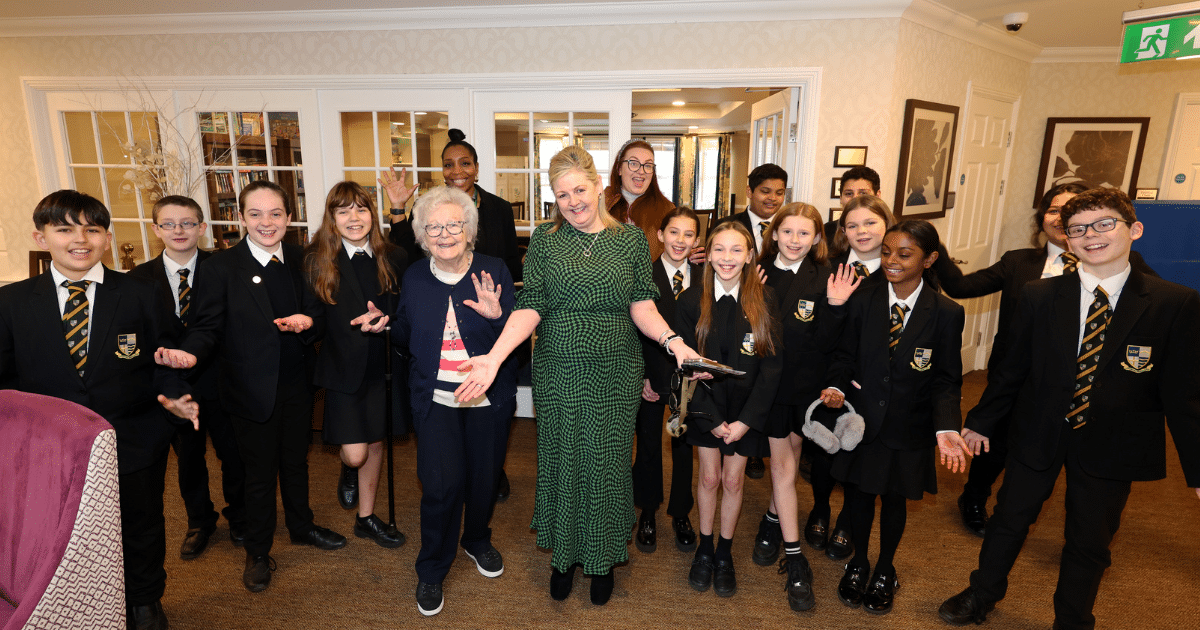 A group of students stand together inside Avery Healthcare residential home, with a resident and home manager, smiling to camera