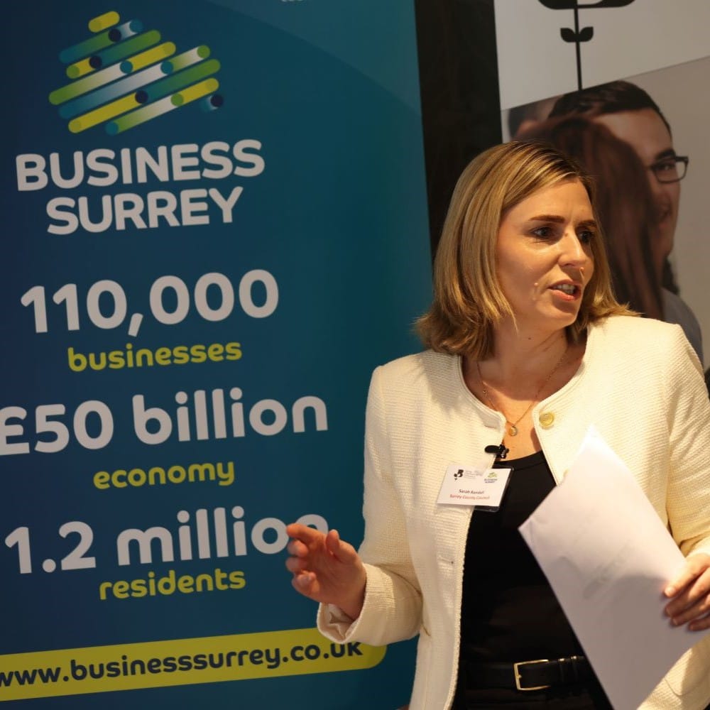 A woman in a white suit jacket holding papers is presenting. She stands in front of a Business Surrey promotional banner.