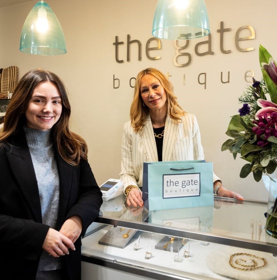 A square image featuring two women smile as they stand either side of a counter in a retail business. Gold lettering spelling The Gate Boutique is on the wall behind them.