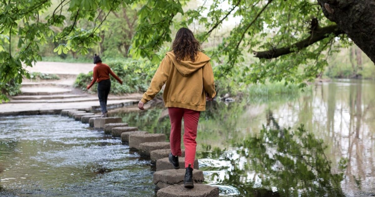 Two women walk carefully across stepping stones in a river. A tree hangs overhead.
