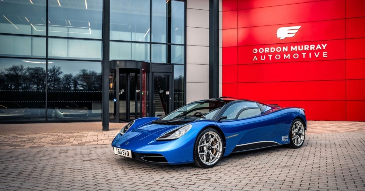 A blue supercar is parked outside a glass fronted office building. On the wall of the building is the logo for Gordon Murray Group