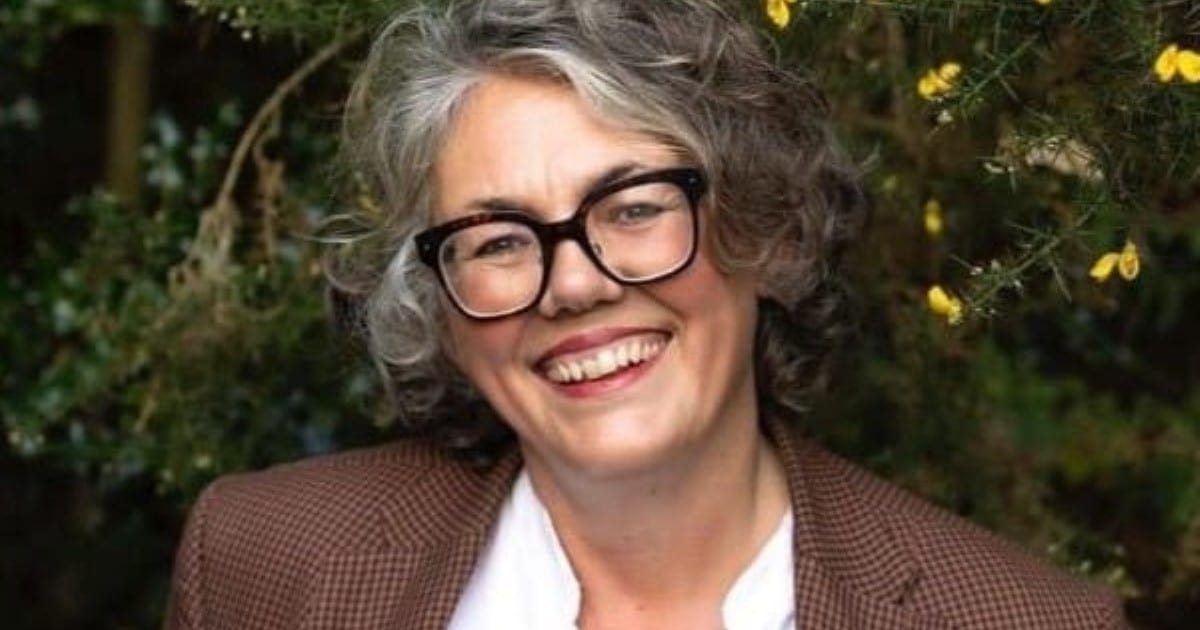 Professional headshot of a smiling woman with black glasses, white shirt and brown jacket. She is standing outside in front of a large green shrub.