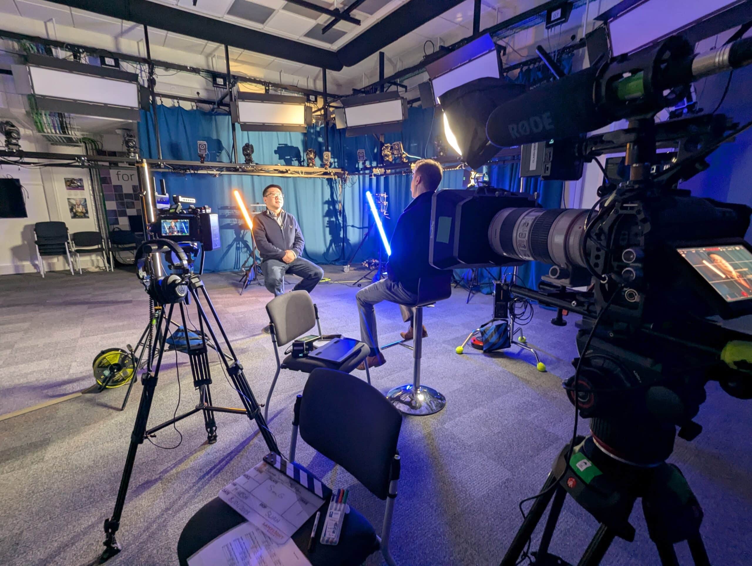 Image of a film set where interviews are taking place behind three cameras. A blue back light is in the background
