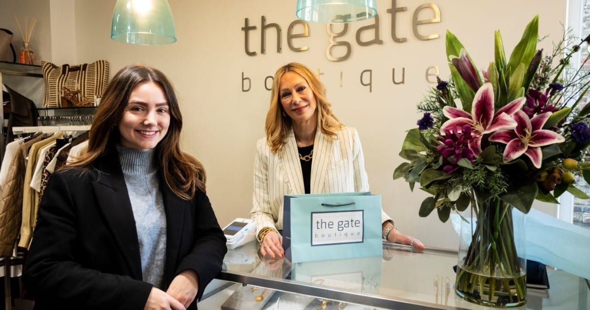 Two women stand in a shop smiling. One is wearing a black jacket and learning on a shop counter, the other is wearing a white jacket and behind the counter. Behind them is a sign for The Gate Boutique, a retail unit in Guildford, Surrey