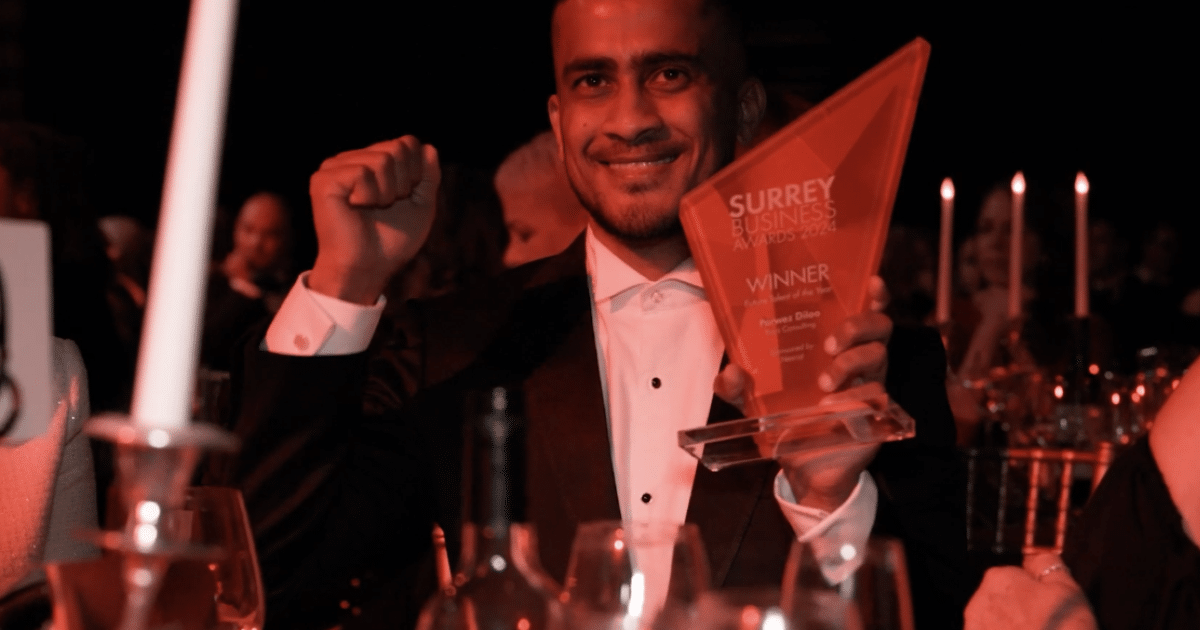 A man in formal suit and white shirt smiles while holding a Surrey Business Awards trophy.