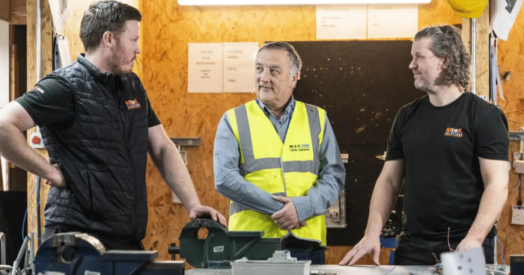 Three men talk to each other in a construction classroom. The man in a middle is wearing a high vis jacket with M.I.T. Skills on it