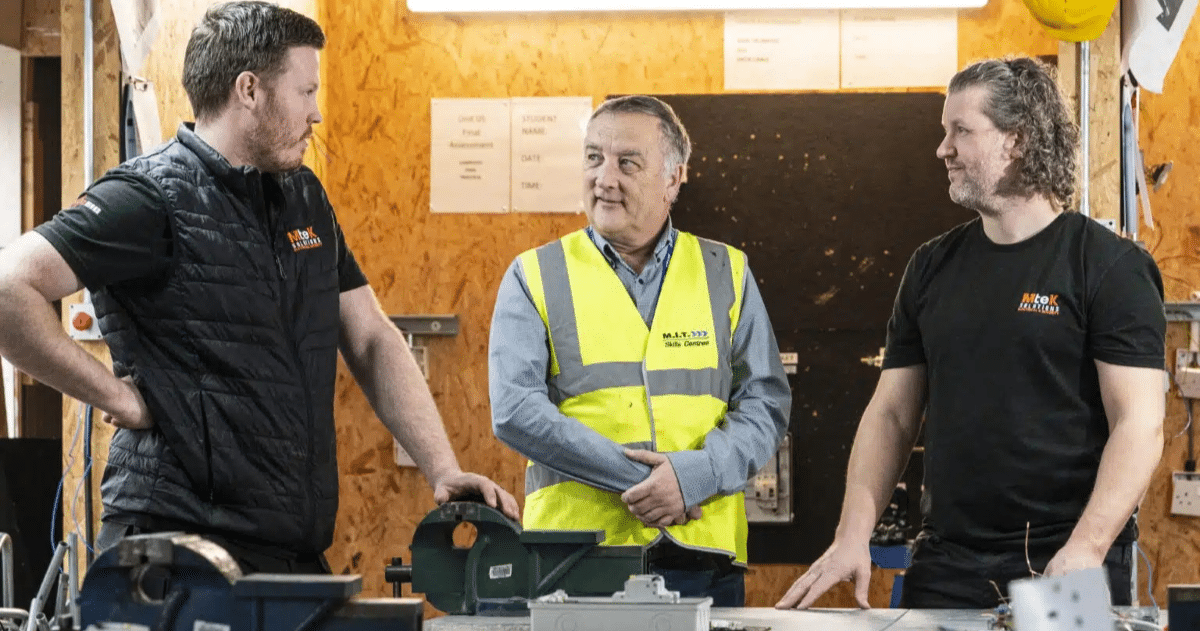 Three men talk to each other in a construction classroom. The man in a middle is wearing a high vis jacket.
