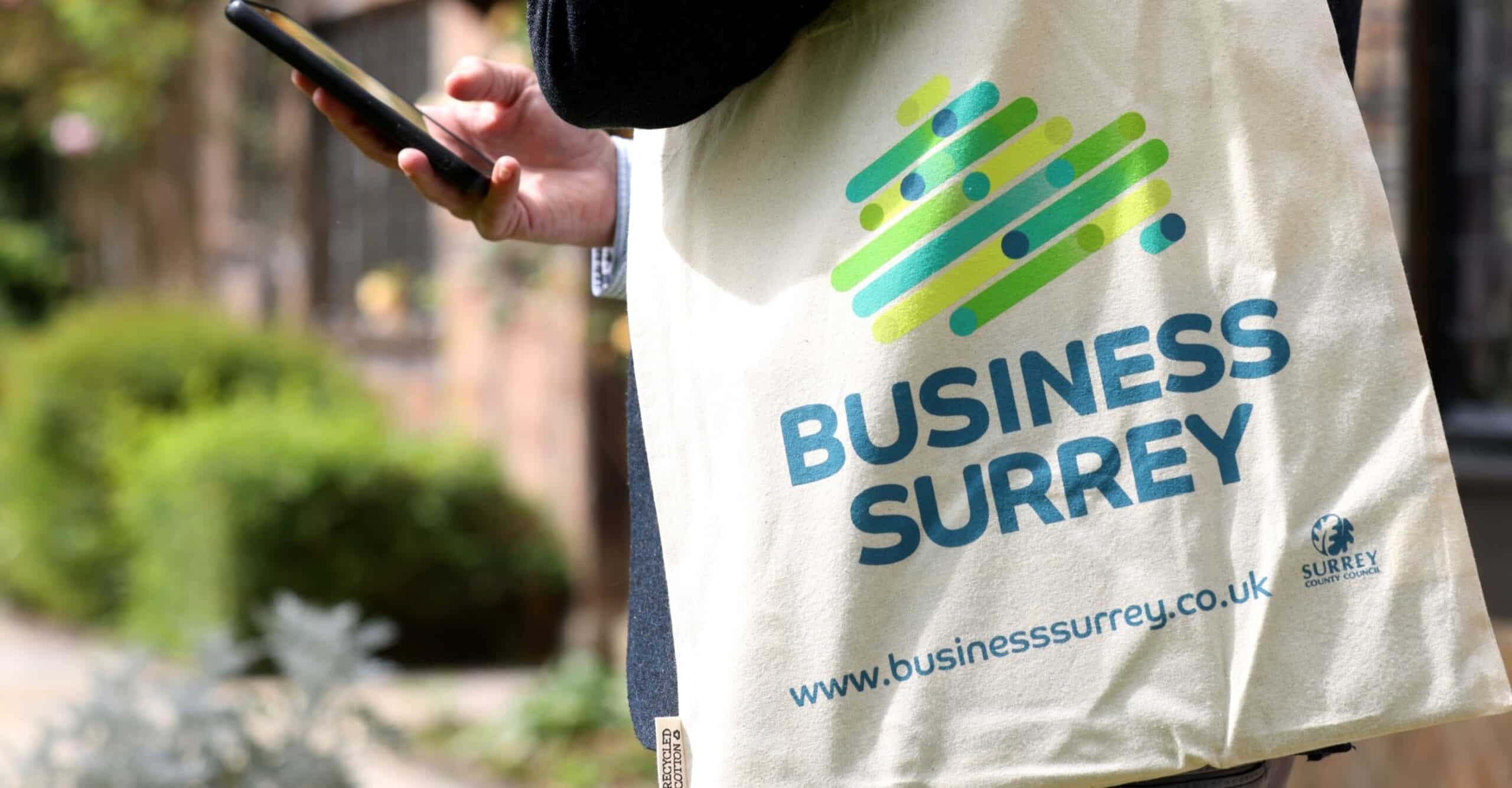 A man holding a tote bag with the Business Surrey logo looks at his phone