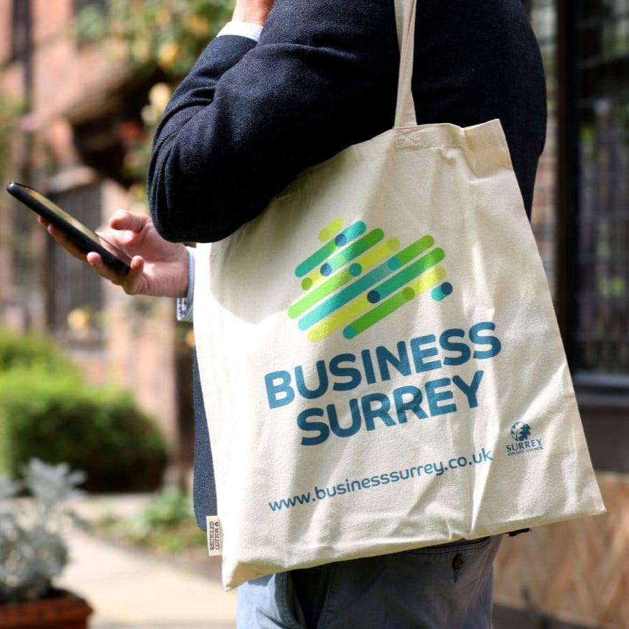 A man holding a tote bag with the Business Surrey logo looks at his phone