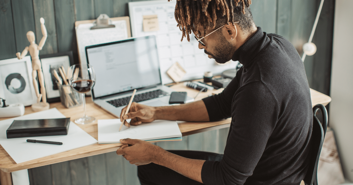 A man with glasses and dreadlocks pulled into a bun sits at a desk writing notes. The desk is full of papers, laptops, screens, etc.