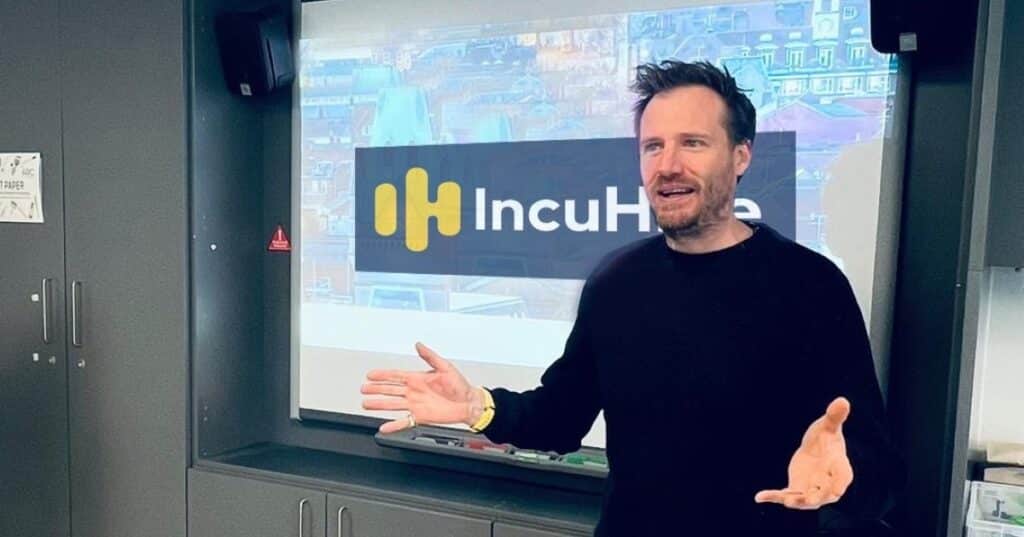 A man in a business setting presents to a room. He is wearing a smart black top and has the words IncuHive written on a screen behind