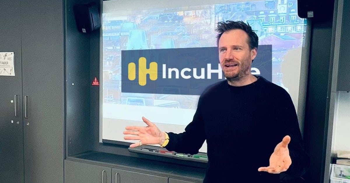 A man in a business setting presents to a room. He is wearing a smart black top and has the words IncuHive written on a screen behind