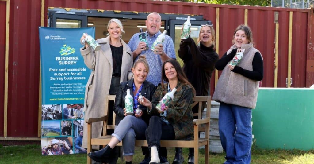 Six people holding craft gin bottles hold them in the air as a toast. Two of the people are sat on a wooden bench. Behind them is a promotional banner for Business Surrey.