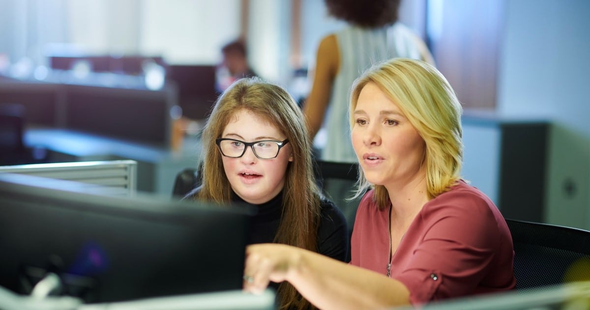 Two women sit together at a computer in an office environment.