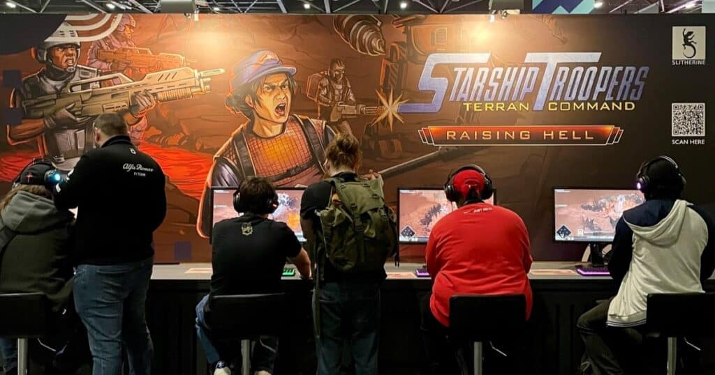 Six people with their backs to the camera are playing video games. Above them is a promotional image for StarShip Troopers, a game by Slitherine Software