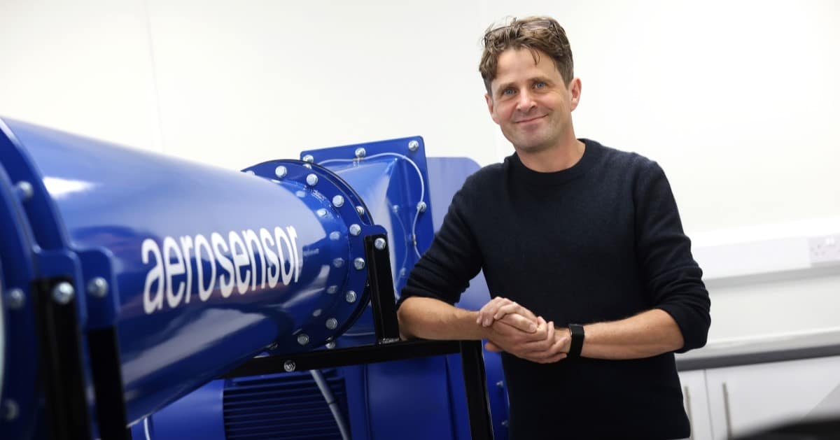 A man in a black jumper stands next to a blue wind tunnel with aerosensor on the side in white lettering