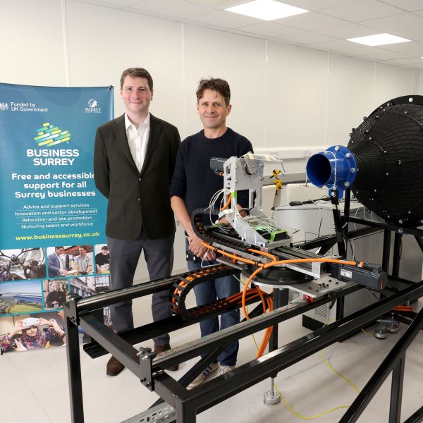 Two men stand next to a high tech wind tunnel in a white warehouse. Behind them is a Business Surrey promo banner