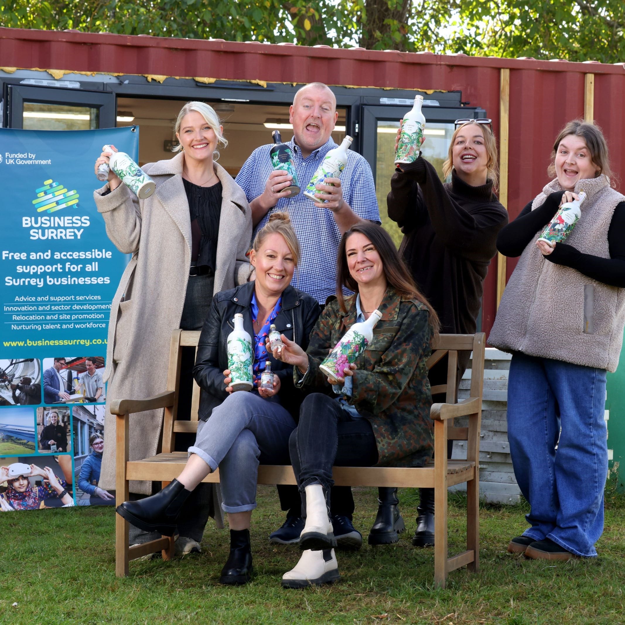 Six people holding bottles celebrate receiving funding. A Business Surrey promo banner is in the background