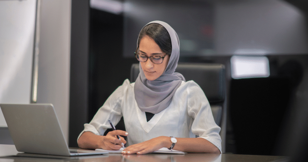 woman sits at desk using a laptop. She is wearing a hijab and dark glasses.