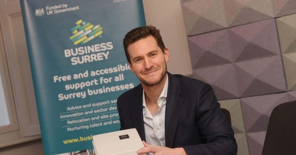 A man in a blue suit and white shirt smiles while posing for an image in front of a Business Surrey promotional banner.