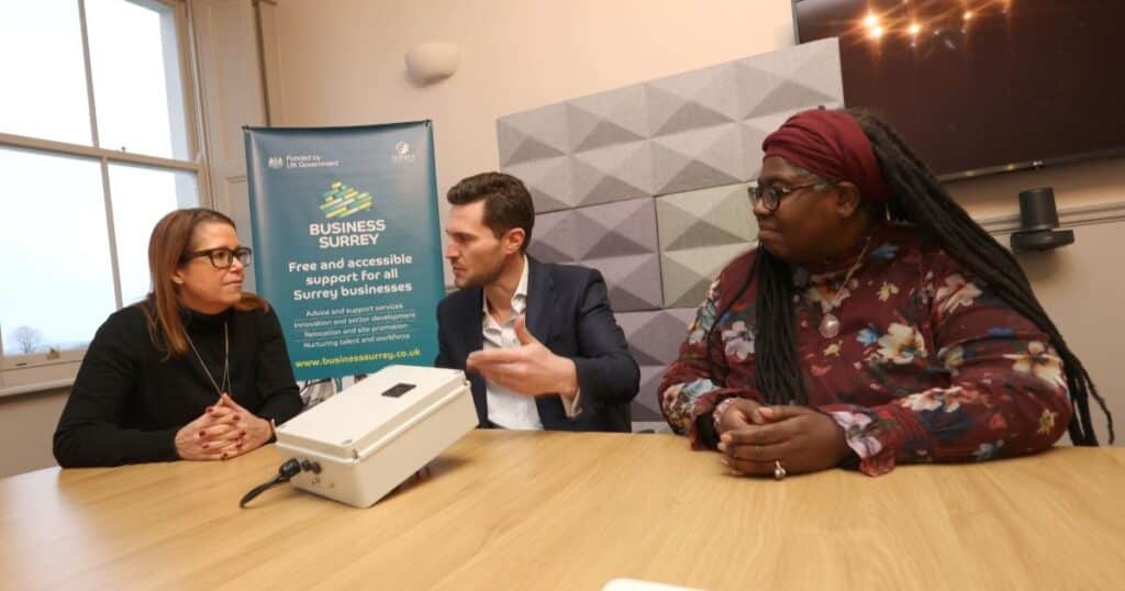 Three people sit at a table discussing a business proposal. Behind them is a Business Surrey promotional banner.