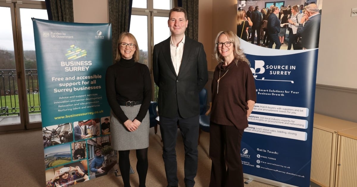 Three people stand smiling in front of promotional banners for Source in Surrey and Business Surrey