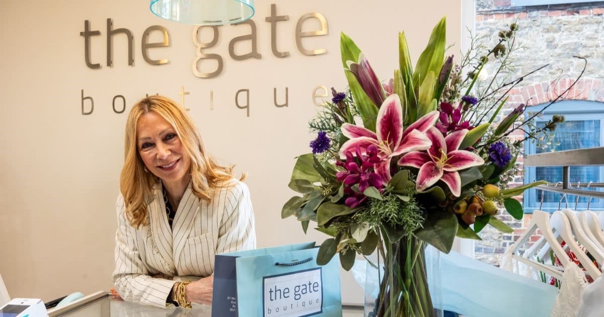 A shop owner of a high street business smiles while stood behind a counter. A large bouquet of flowers stand in a vase next to her.