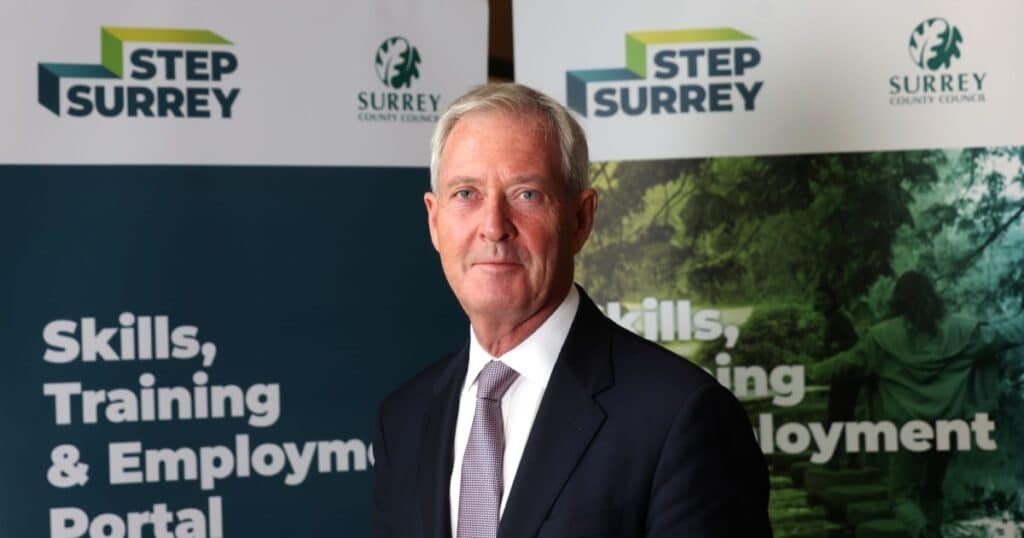 A man in a black suit, white shirt and tie stands in front of two promotional banners for STEP Surrey | Business Surrey