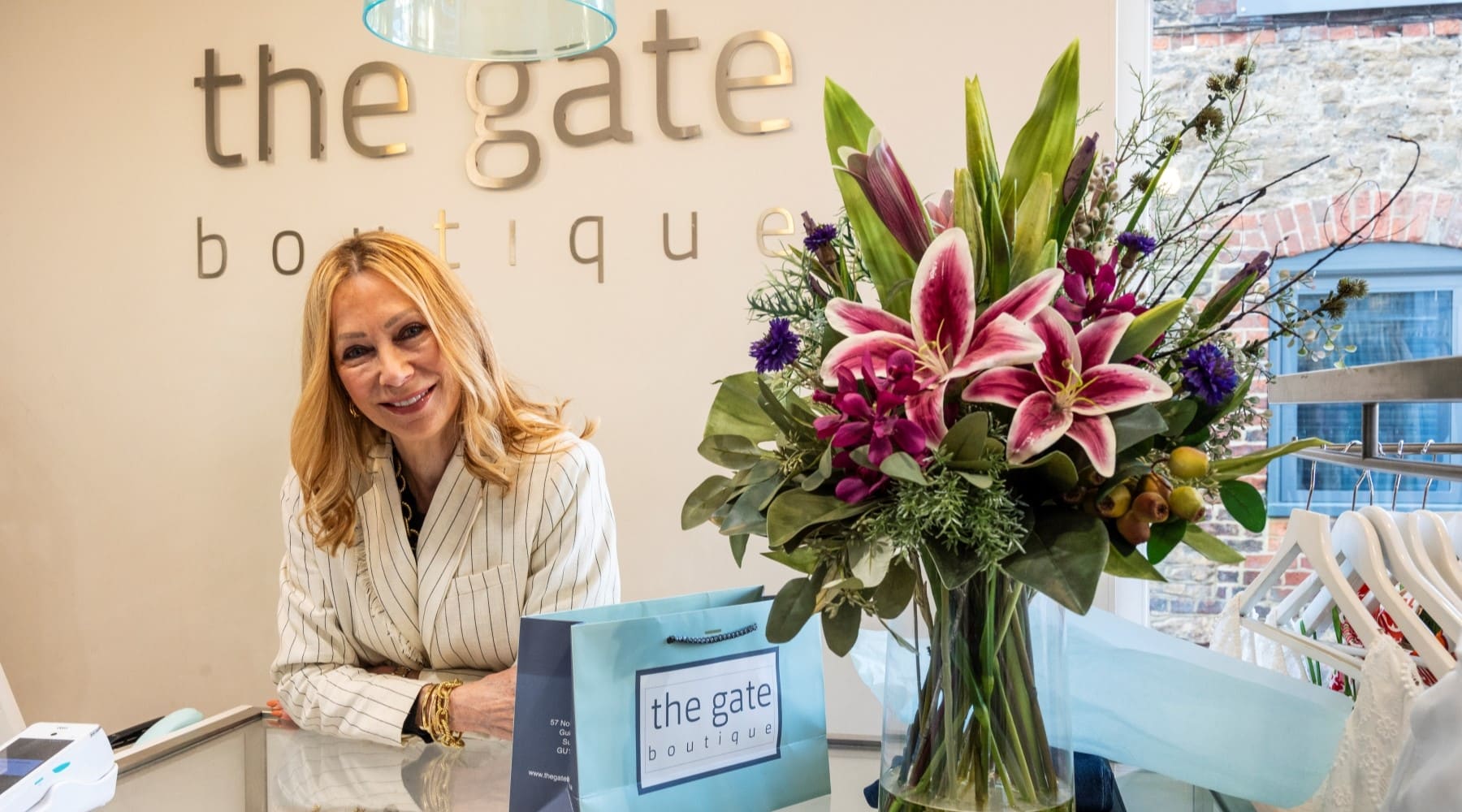A shop owner of a high street business smiles while stood behind a counter. A large bouquet of flowers stand in a vase next to her.