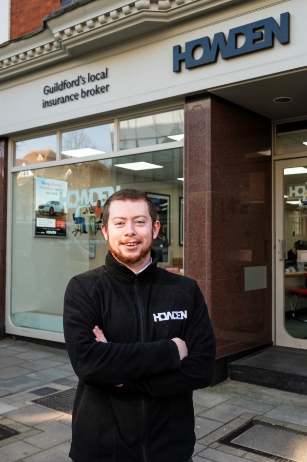 A man in a black fleece with Howdens on it stands with his arms folded in front of a high street business.