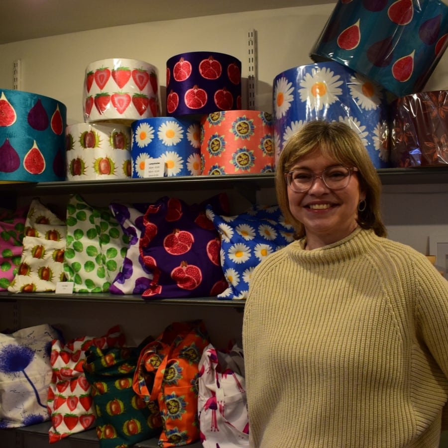 A shop owner in a cream jumper smiles at the camera. Behind her are a series of bright designed goods.