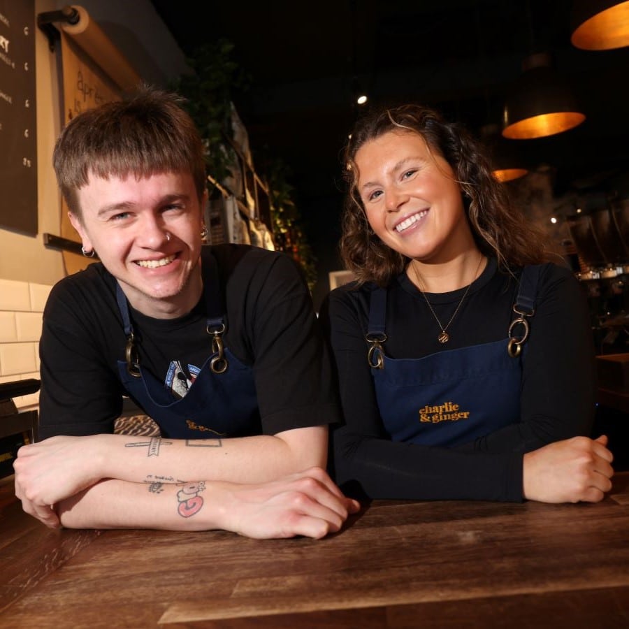 Two coffee shop workers in blue aprons stand smiling behind the counter