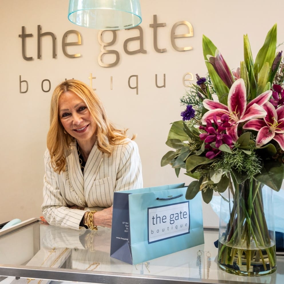 Square image of a shop owner of a high street business smiles while stood behind a counter. A large bouquet of flowers stand in a vase next to her.
