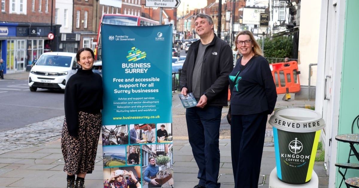 Three people stand posing in a busy high street next to a promotional banner for Surrey County Council's Business Surrey service.