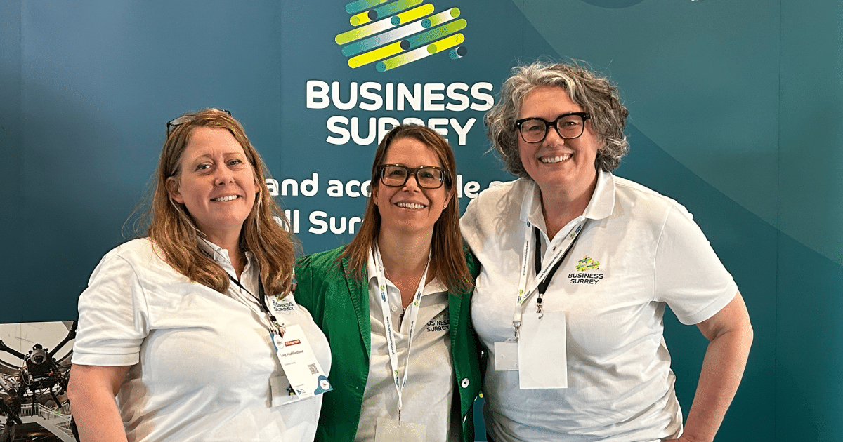 Three women stand in front of a Business Surrey exhibition stand, smiling at the camera.