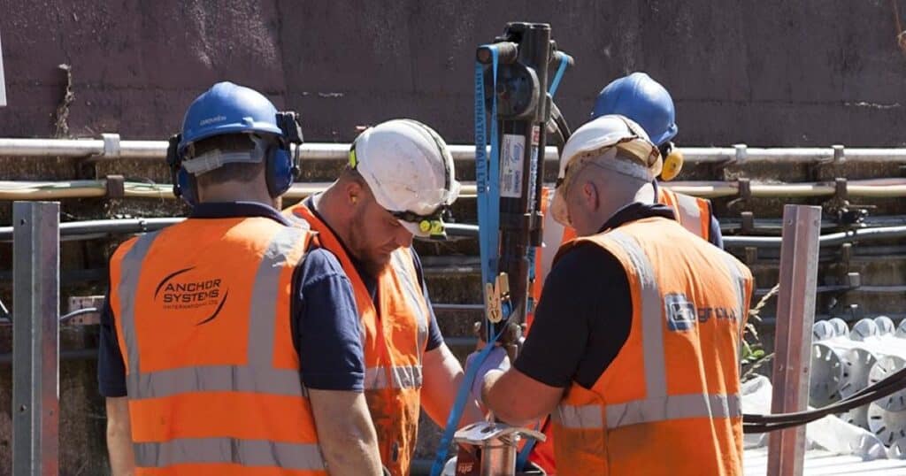 Four people in high vis vests with Anchor Systems on the back stand around a piece of high tech machinery which is on a construction site