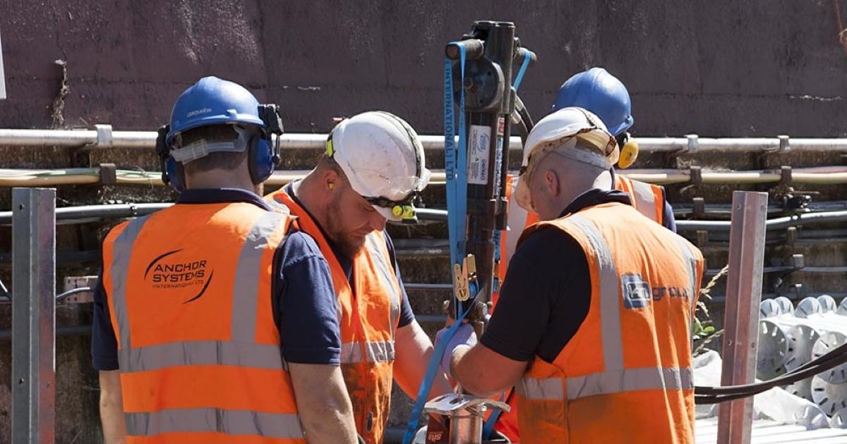 Four people in high vis vests with Anchor Systems on the back stand around a piece of high tech machinery which is on a construction site