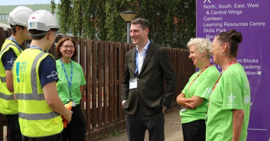 A man in a black suit stands talking to young people wearing high vis at a further education college in Surrey