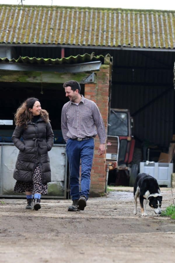 Upright image of two people walking and talking while on a farm in Surrey. Behind them is a barn and a parked tractor while a sheep dog walks by the farmer's feet.