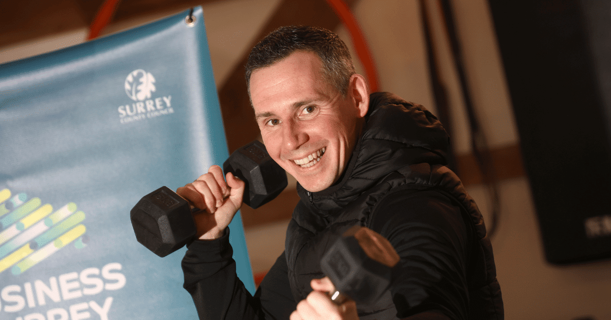 Frank holds two dumbells in front of a Business Surrey banner and smiles at the camera | Fit with Frank | Business Surrey