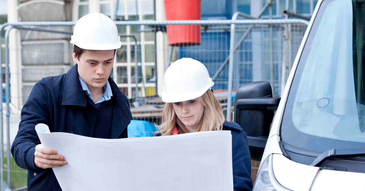 a man and woman wear hard hats on a construction site. they are reading a large sheet of paper - plans. | Business Surrey | South East business funding